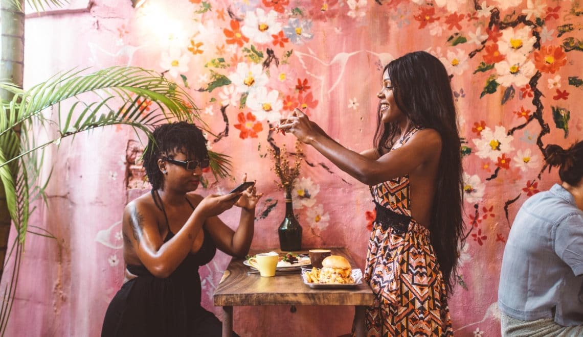 Two Black women take photos of their food with a beauitful floral wall behind them